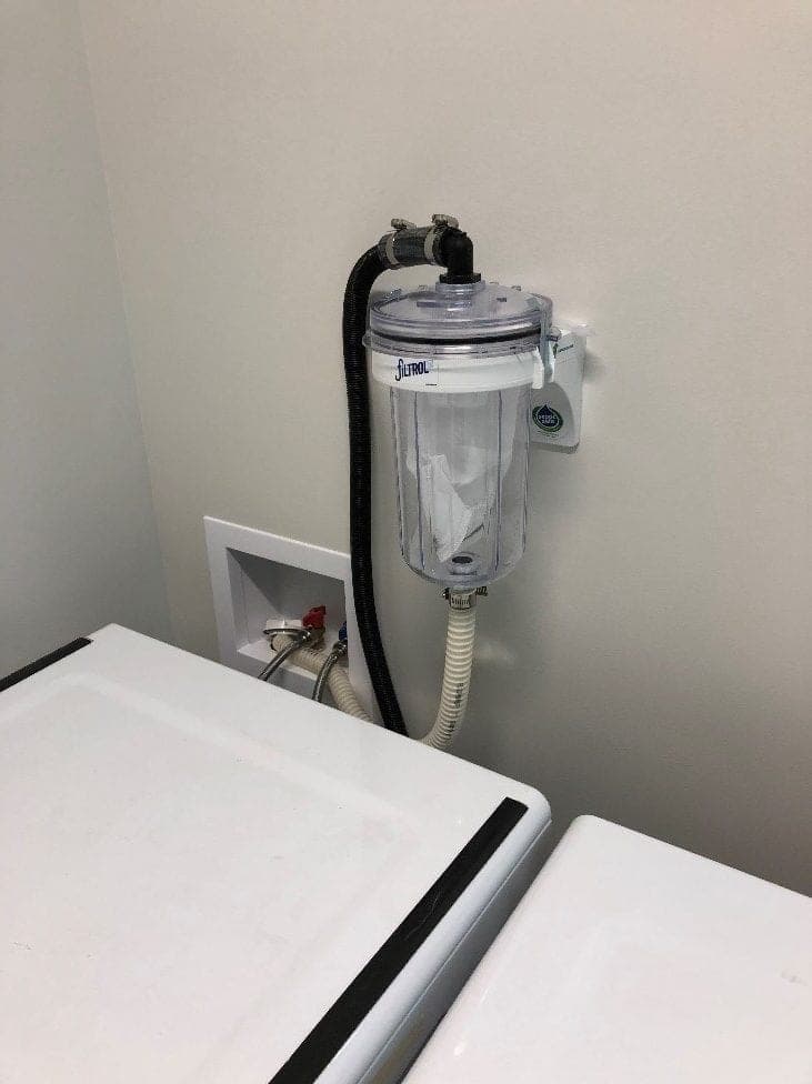 A laundry filter mounted on the wall beside a washing machine, its hose clamped to the machine's discharge line above the wall box