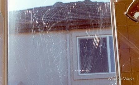 Long arcing scratch tracks in tempered glass, left by fabricating debris dragging under a blade.