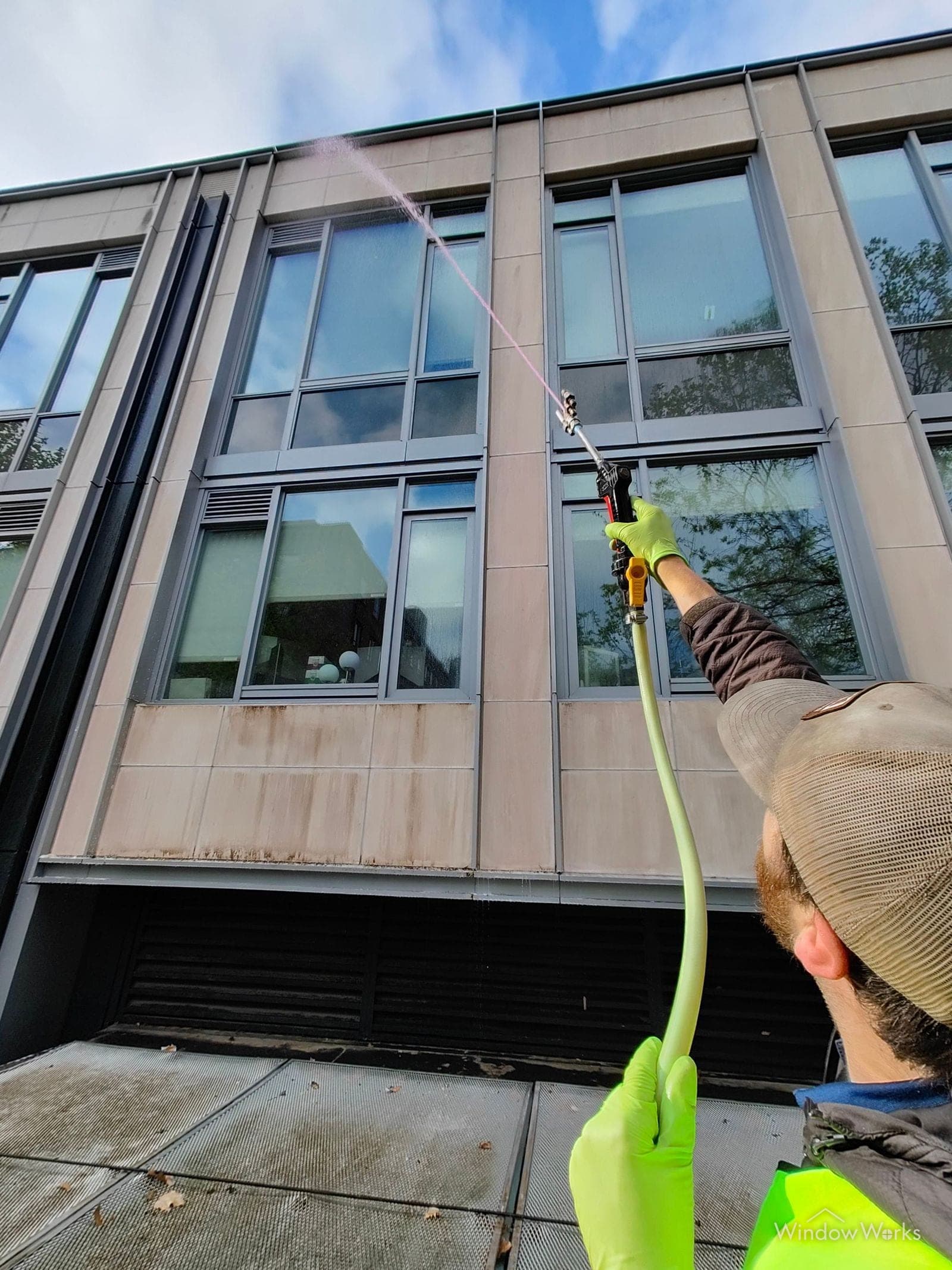 A WindowWorks technician working a wand across the brown-stained stone and glass of a building facade, the clean line following the spray