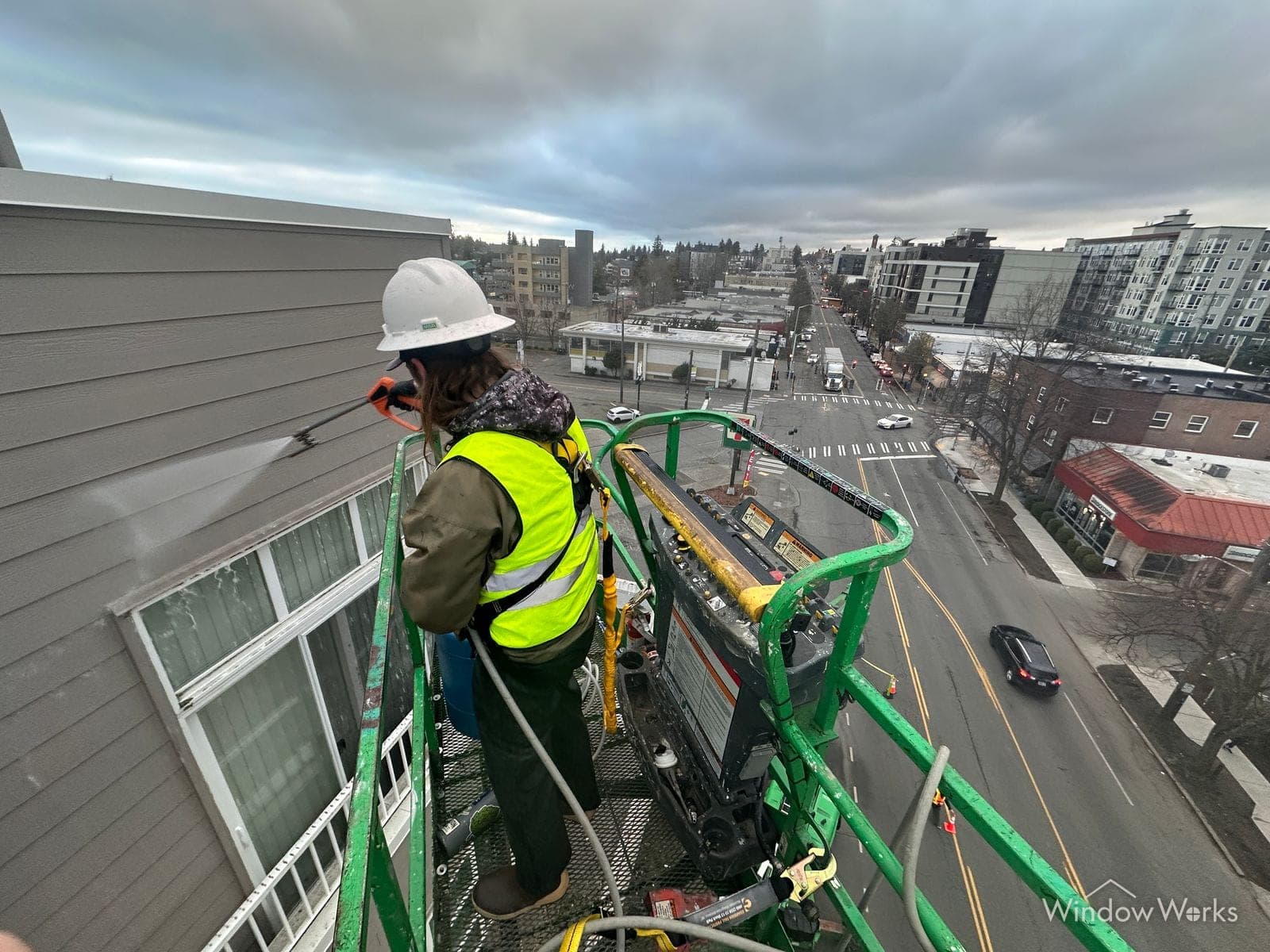 A WindowWorks technician in a hard hat and safety harness washing the upper siding of a building from a boom lift basket, the street and rooftops of Seattle far below