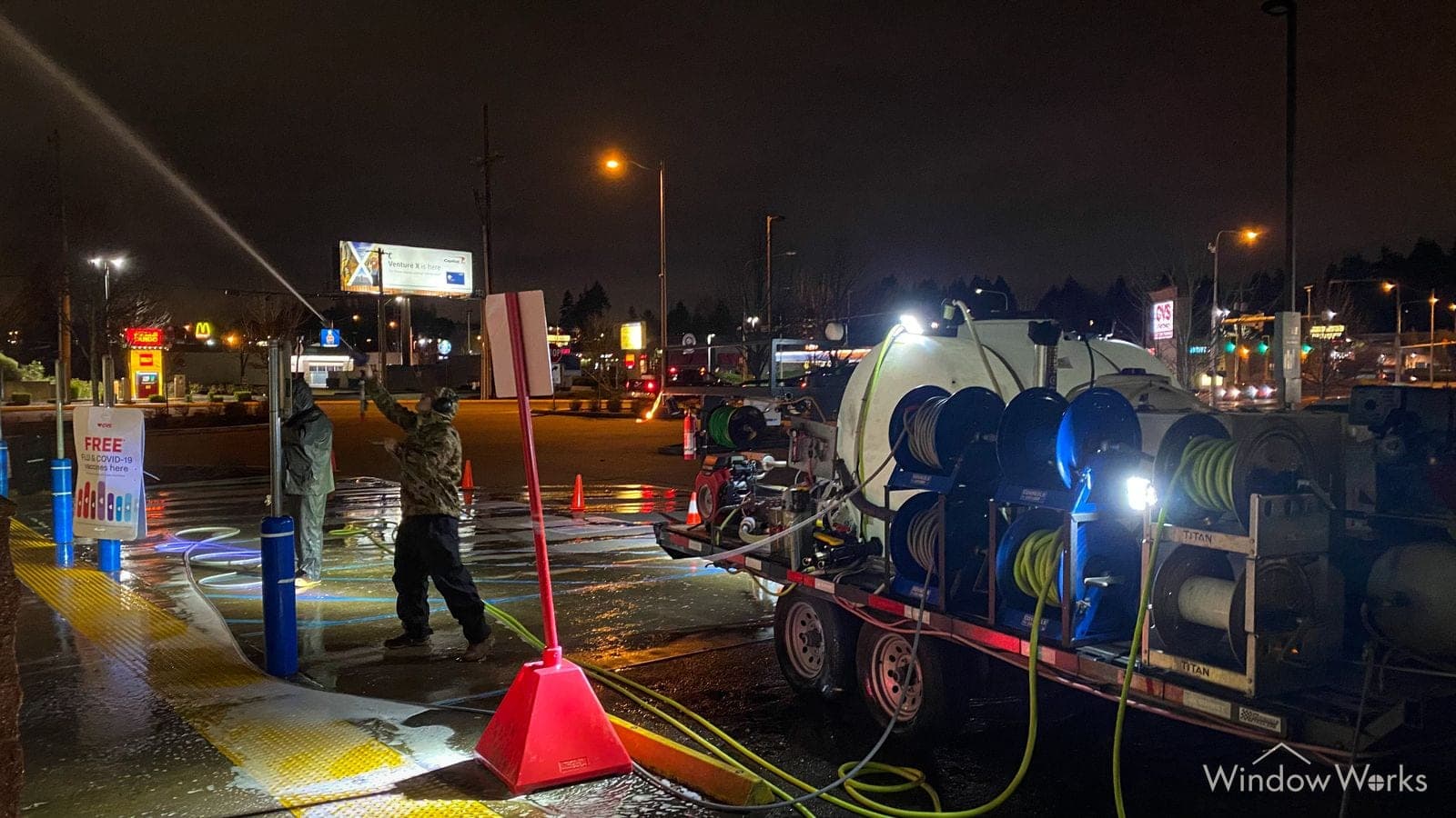 A WindowWorks technician washing a commercial lot at night beside the hot-water rig, cones and hose runs laid out across the wet asphalt