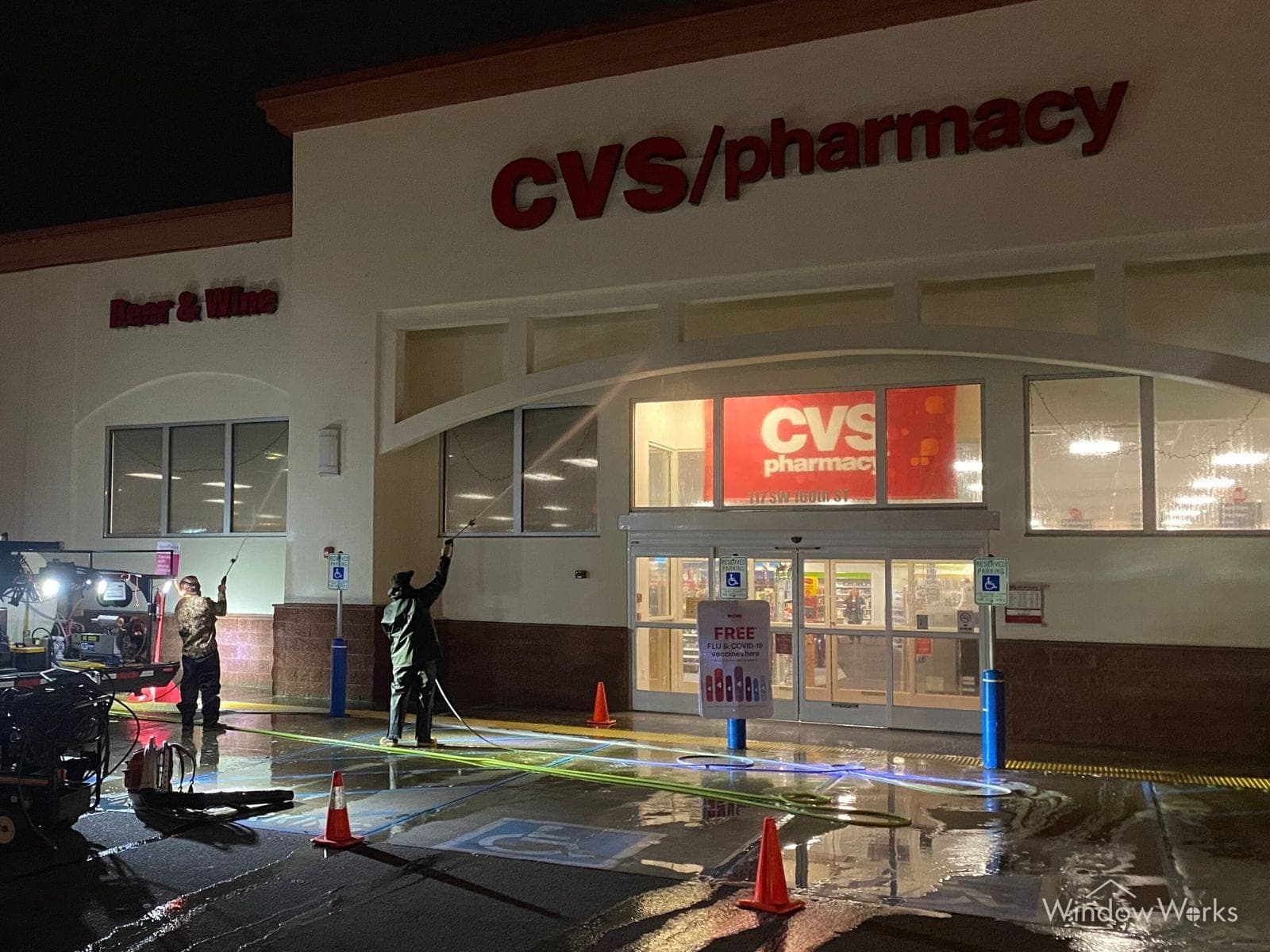 Two WindowWorks technicians washing the front of a closed CVS pharmacy at night, the pressure rig running and traffic cones set out across the wet lot, on the Eastside of Seattle