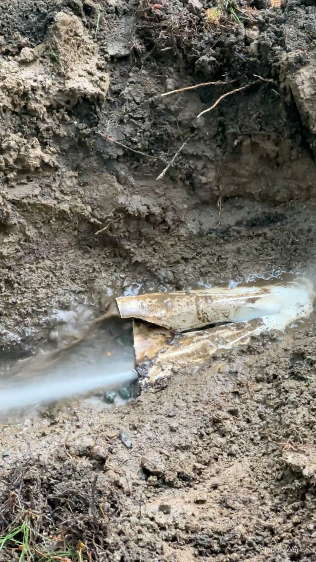 A crushed and split underground drain pipe exposed in an excavated hole, water blasting clear through it after jetting