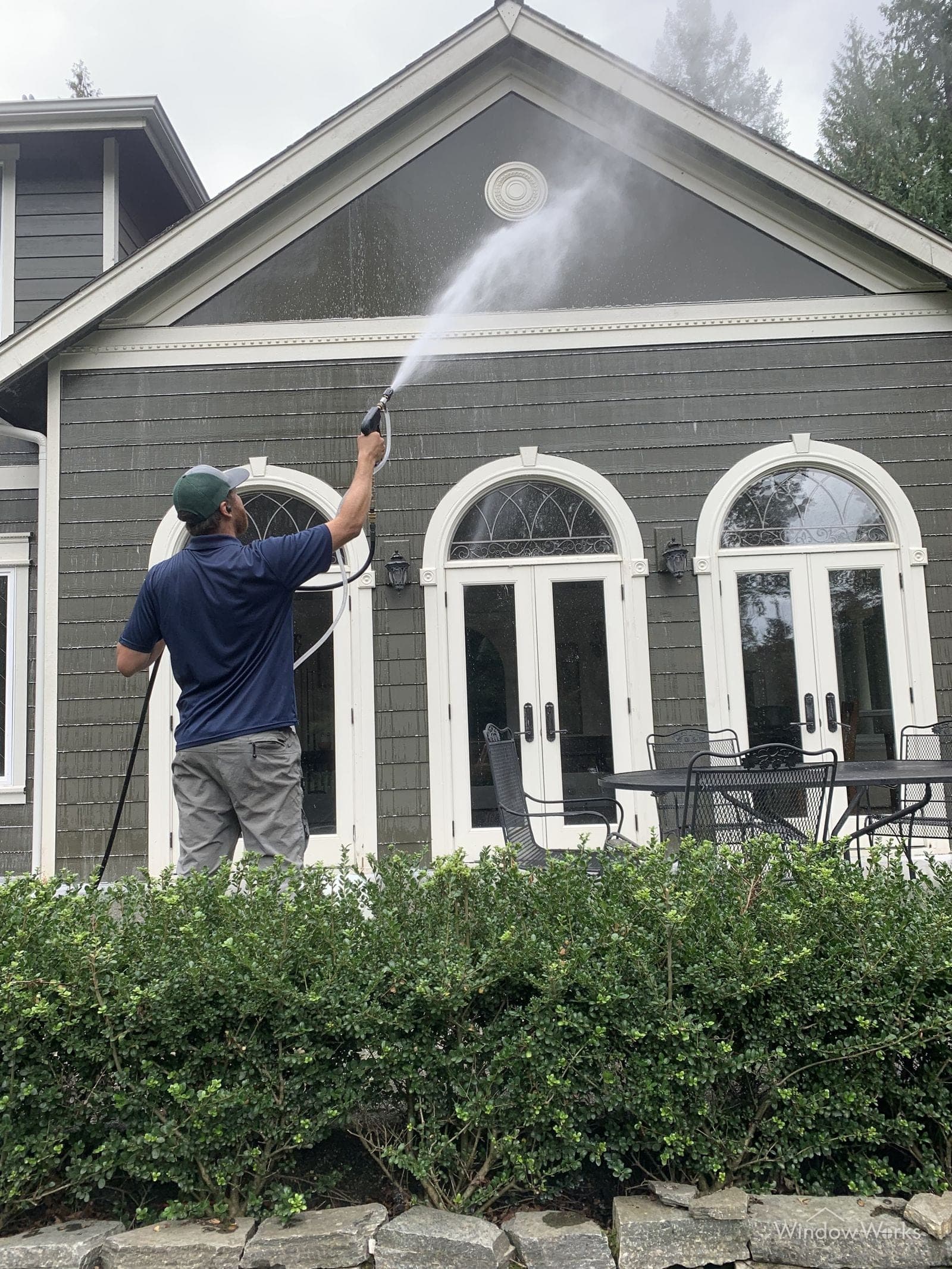 A WindowWorks technician soft washing the gable of a gray shingle-sided home above its arched French doors on the Eastside