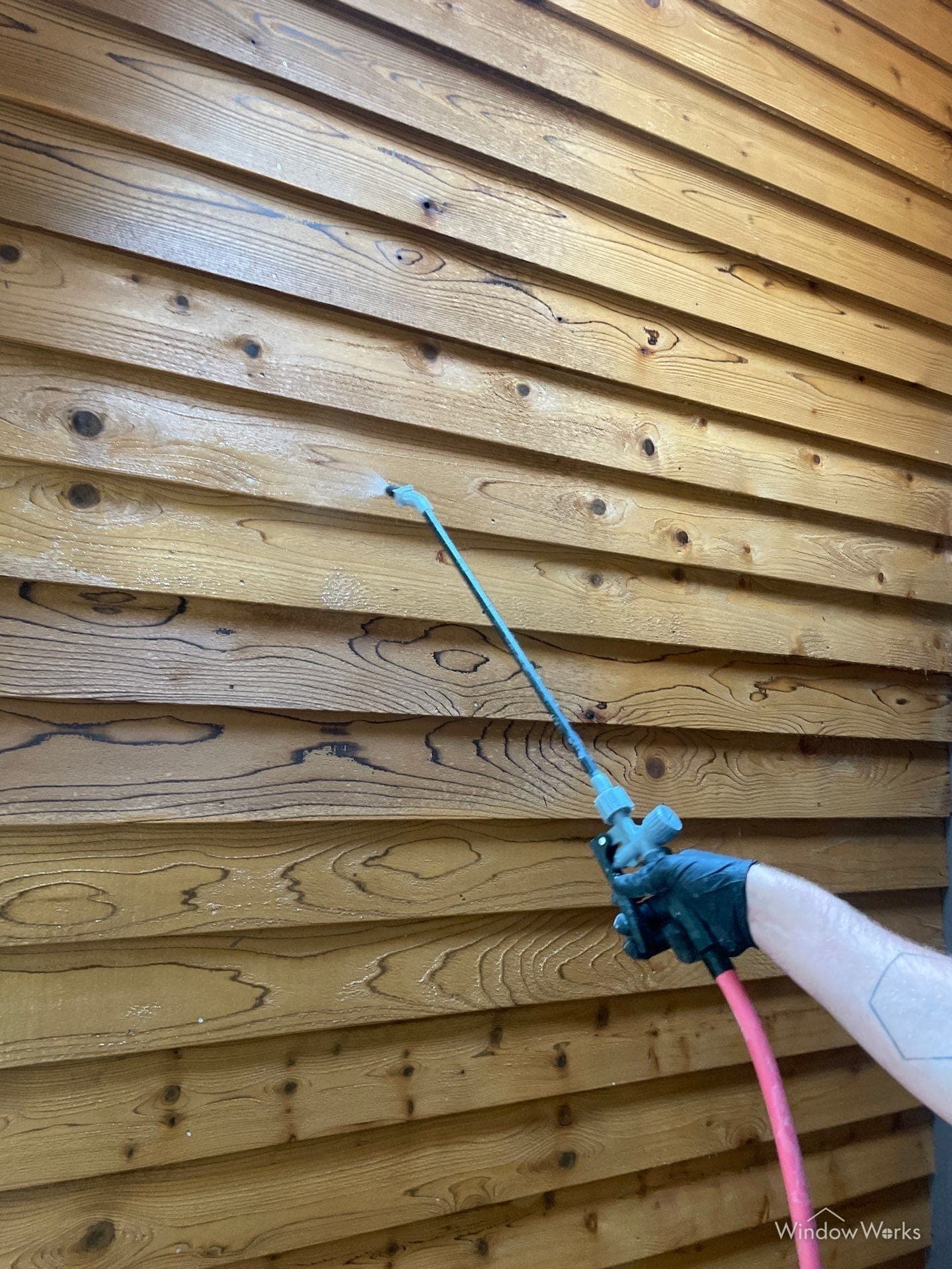 A gloved WindowWorks technician spraying oxalic acid brightener onto tannin-darkened cedar boards on the Eastside
