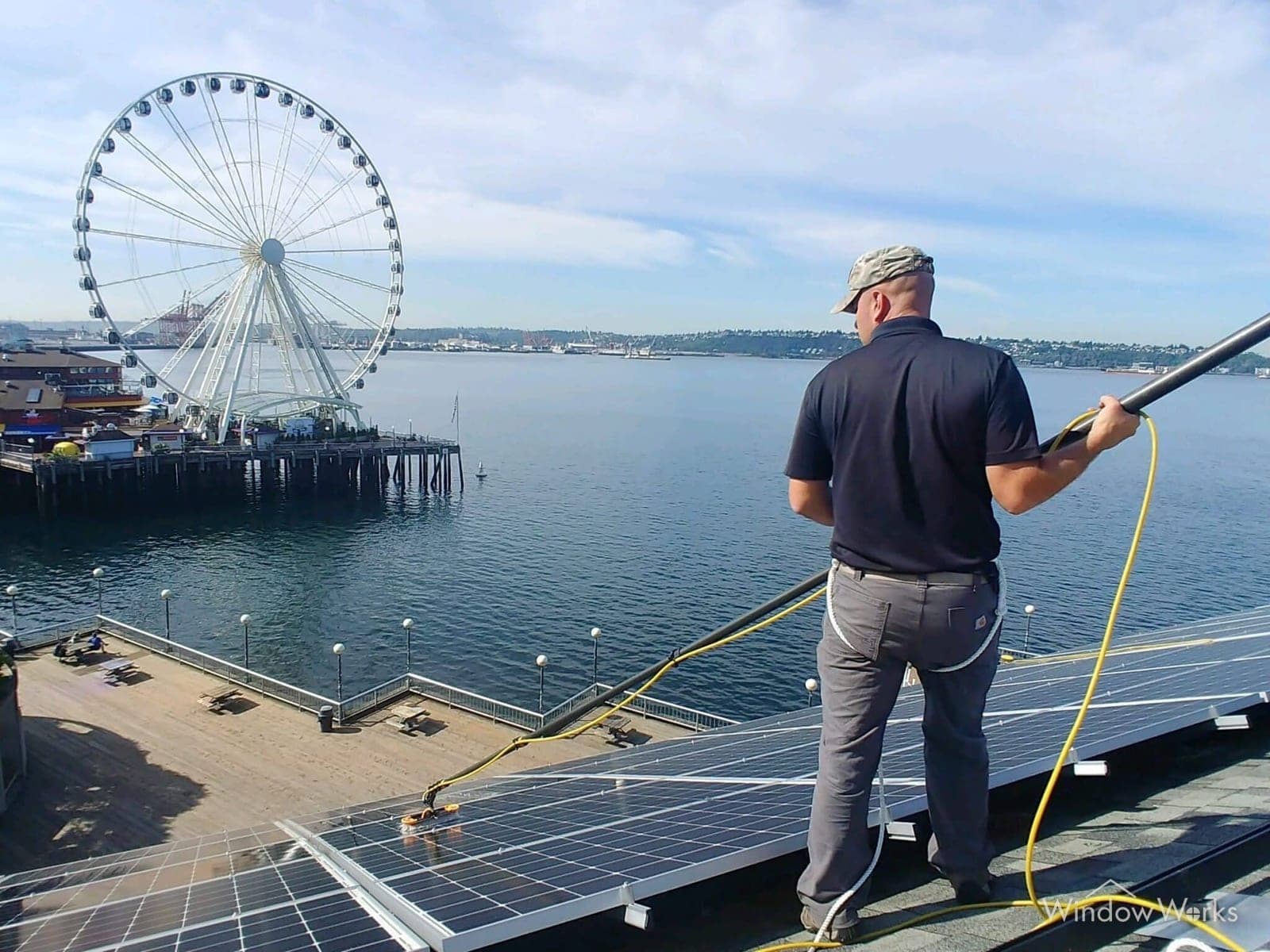 A WindowWorks technician cleaning rooftop solar panels with a DI-water-fed pole above the Seattle waterfront on the Eastside