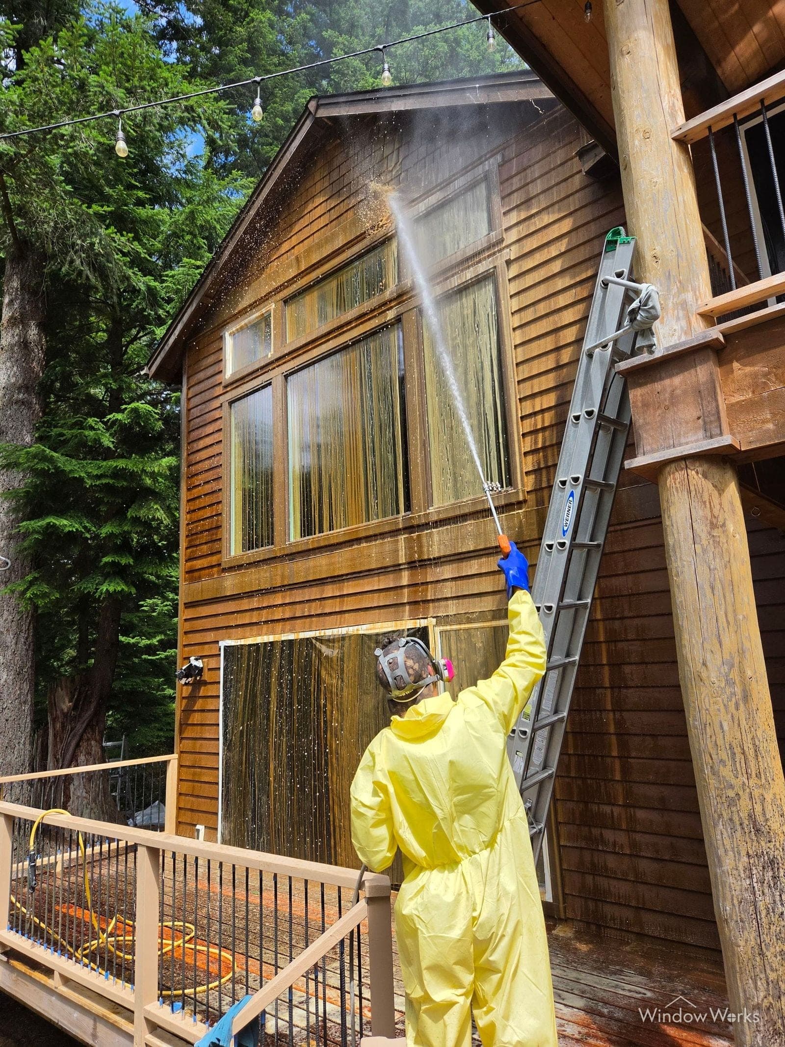 A WindowWorks technician in a full protective suit spraying chemical stripper up a cedar gable, the old finish already running off the masked wall