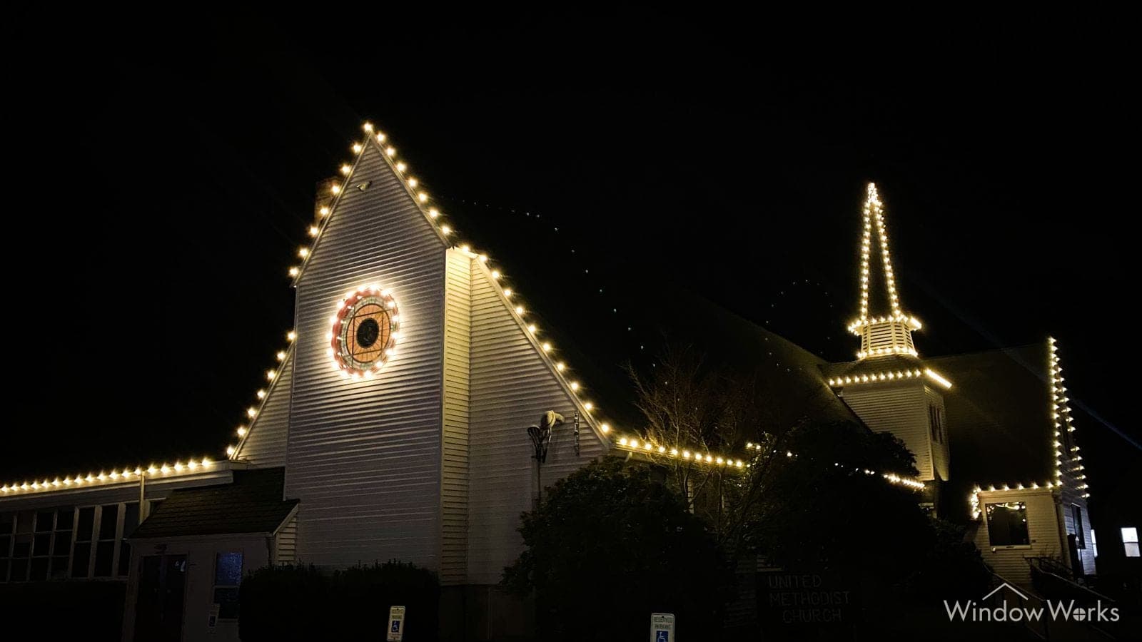 A church at night with warm white C9 lights tracing its steep gable and steeple, and a lit wreath centered in the round window.
