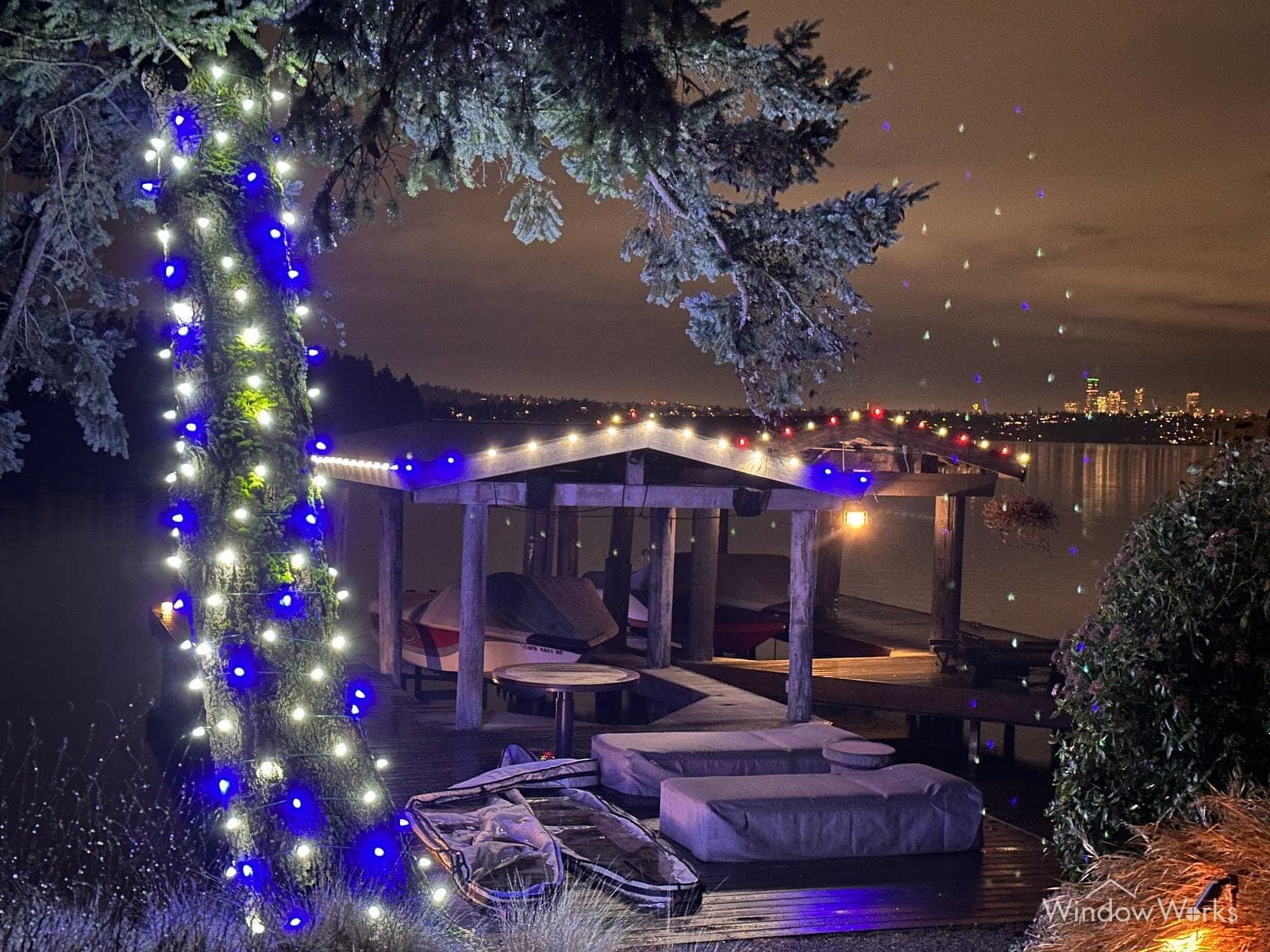 A lakefront boat dock at night, its posts wrapped in blue and white lit garland with the water and far shore lights behind.