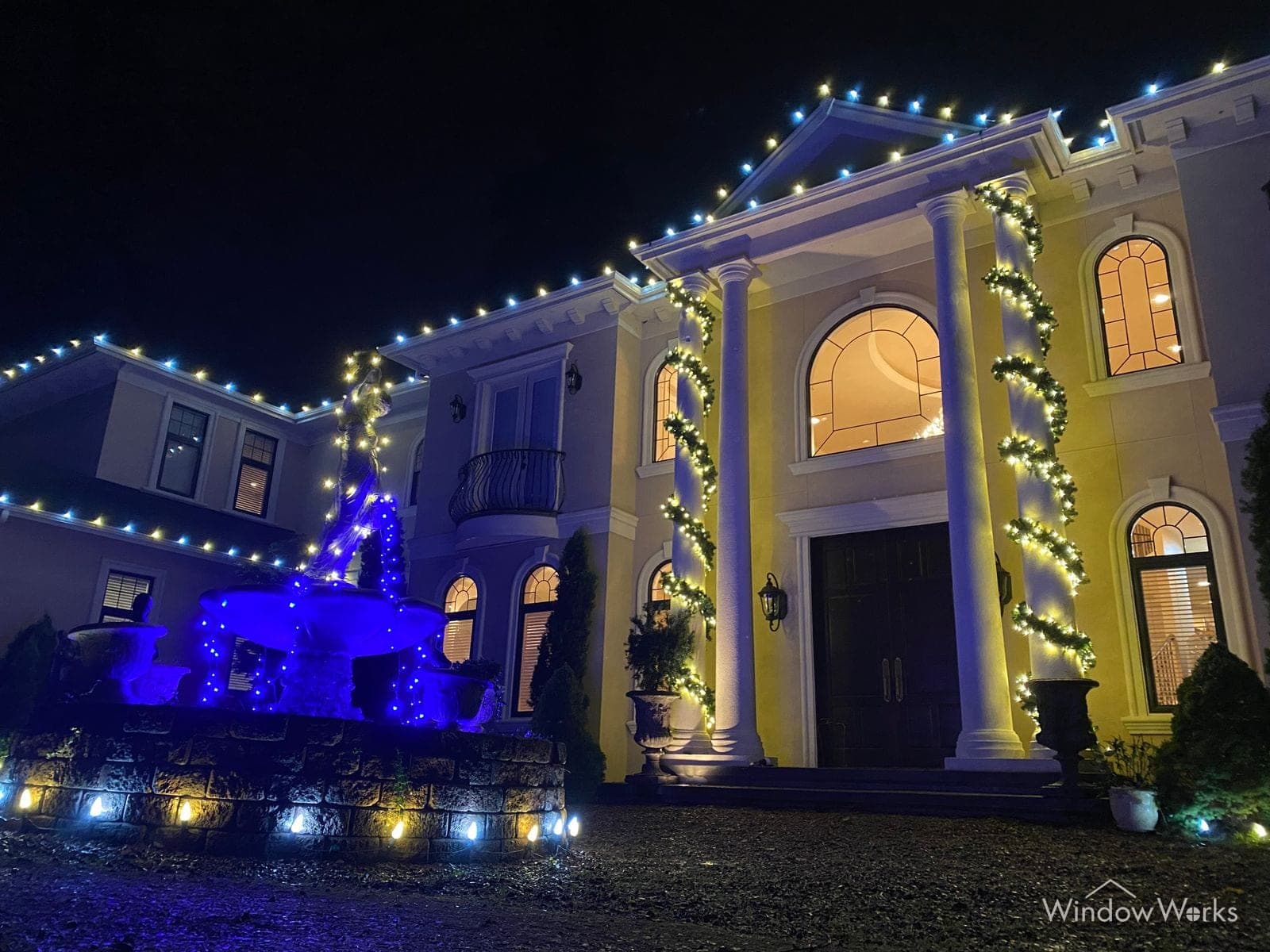 A pillared estate at night with warm white rooflines, lit garland wrapping the columns, and blue accent lighting by the entry.