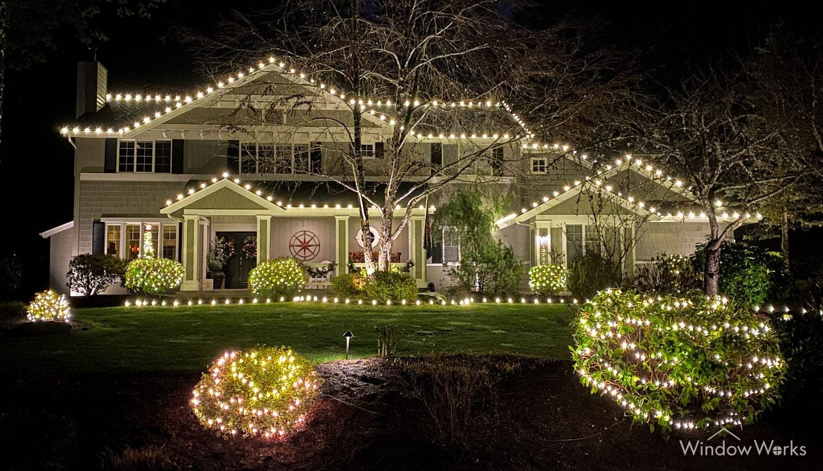 A large Eastside home at night, every roofline traced in warm white C9 bulbs, with lit shrubs, garlands and window wreaths across the front yard.