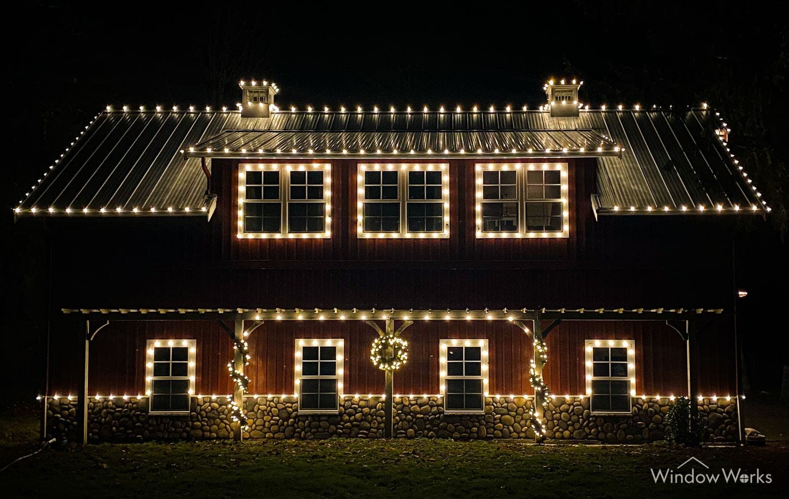 A dark timber carriage barn with warm white bulbs along the roofline, around every window and down the porch posts, wreaths hung between.