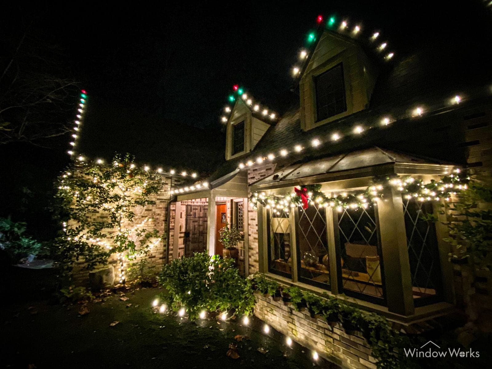 A cottage lit warm white along the eaves with green and red bulbs at the roof peaks and a run of path lights out to the lawn.