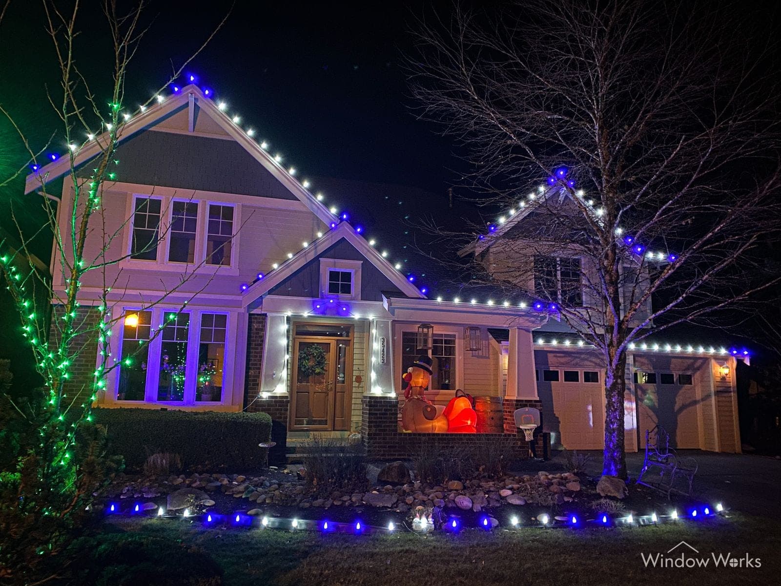 A craftsman home at night in blue and white bulbs, the trees either side wrapped to match and a run of bulbs edging the lawn.