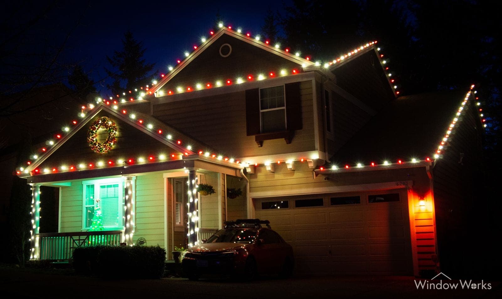 A craftsman home traced in red and green bulbs with a lit wreath in the gable and the porch washed green behind.