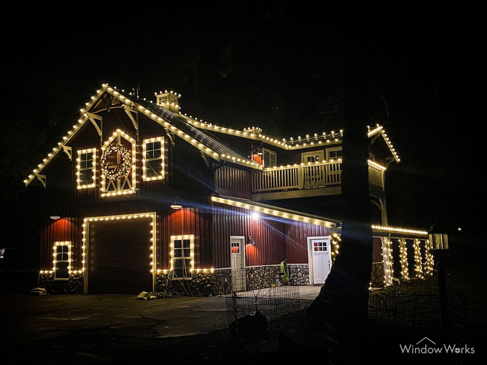 A craftsman home outlined in warm white bulbs with a lit wreath in the gable, the garage and doors framed in bulbs.