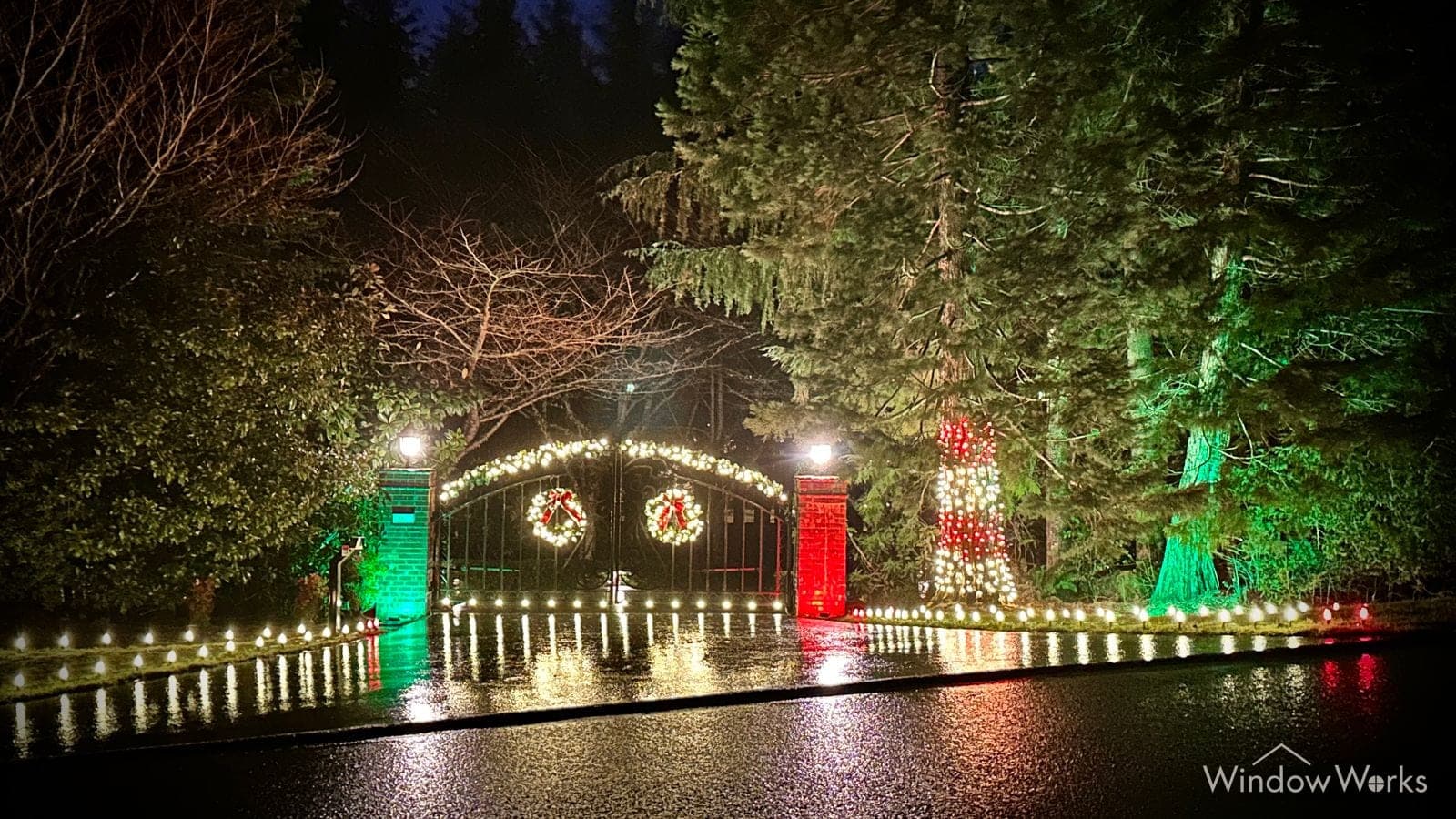A wide view of a lit estate gate with wreaths, warm white bulbs along the roadside wall and firs uplit green and red behind, all mirrored in wet asphalt.