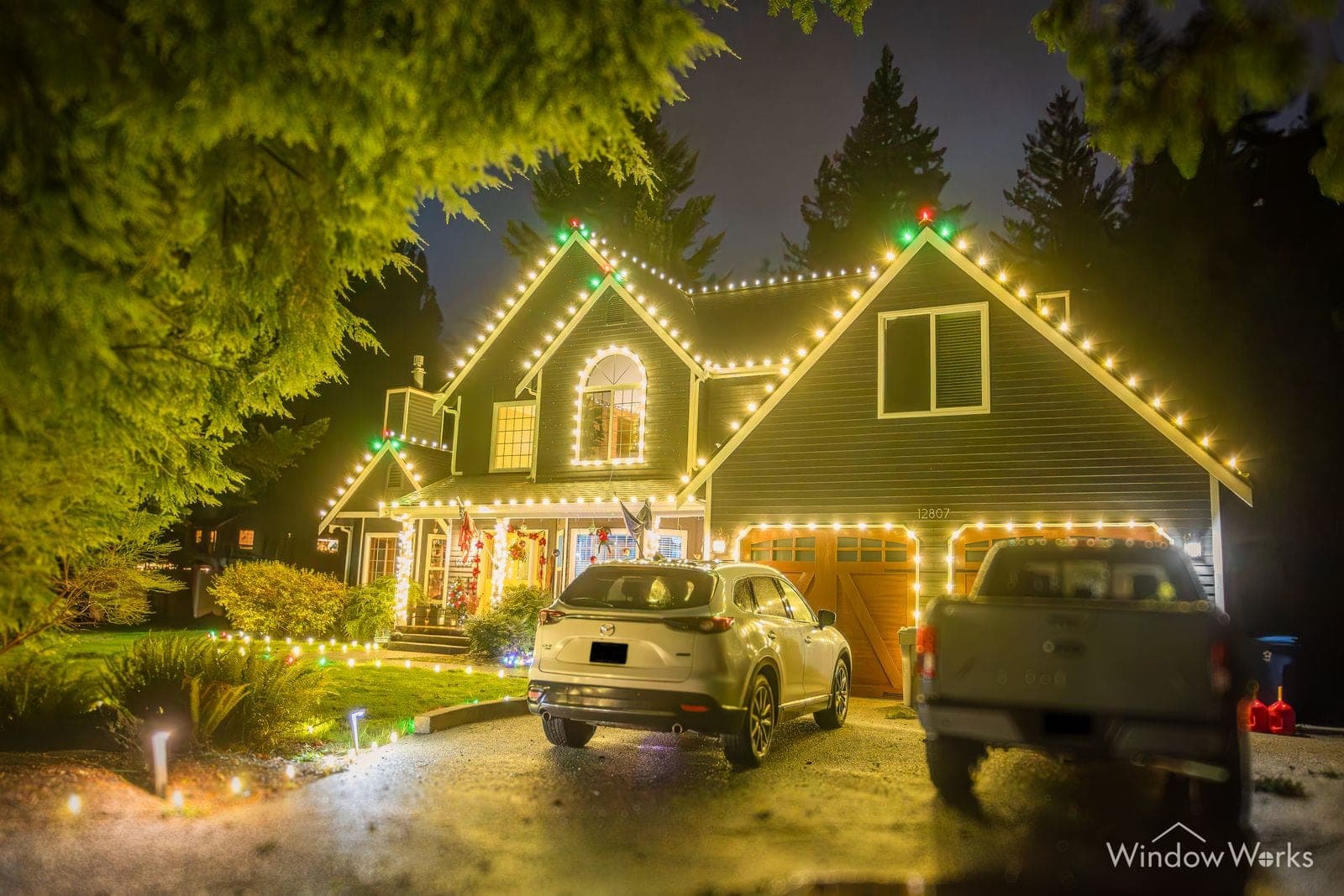 A cedar-shingled home in warm white bulbs with wreaths on the gables and two vehicles on the wet drive.