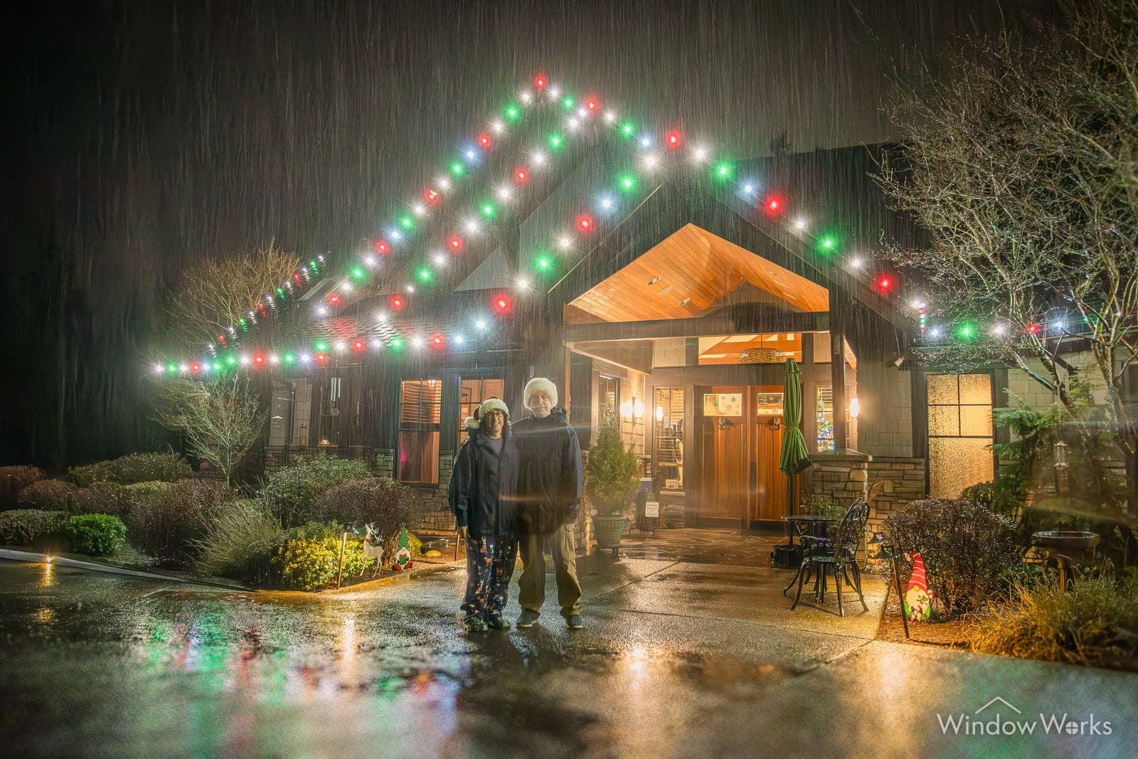 A shingled entry in red, green and blue bulbs with two people at the door on a wet night.