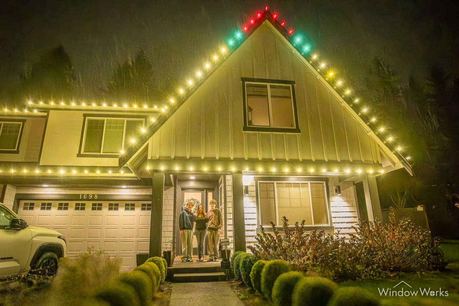 A modern farmhouse in warm white with green and red at the peaks and two people at the lit entry.