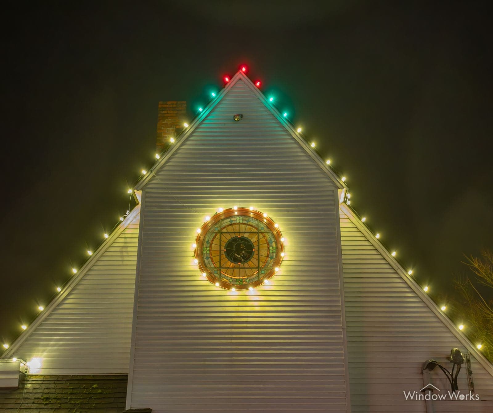 A white gable outlined in warm white bulbs with a large lit wreath at its center.