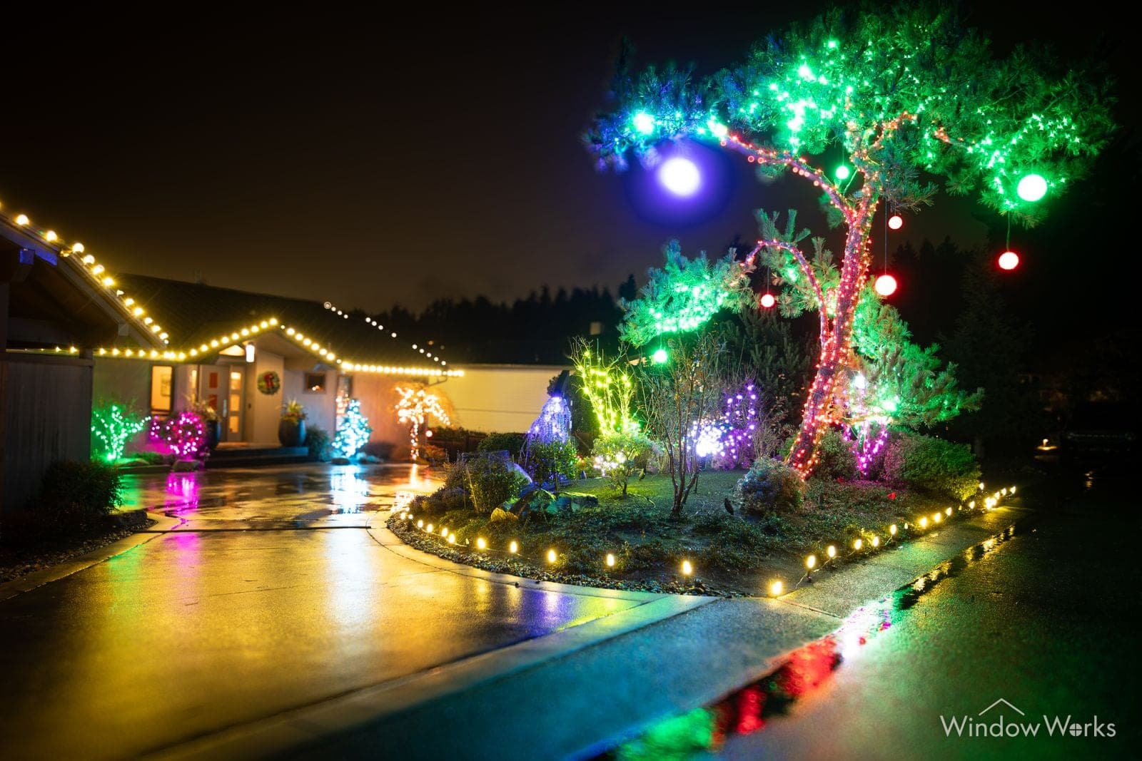 A green-lit tree hung with colored baubles over a lit drive, warm bulbs edging the beds.