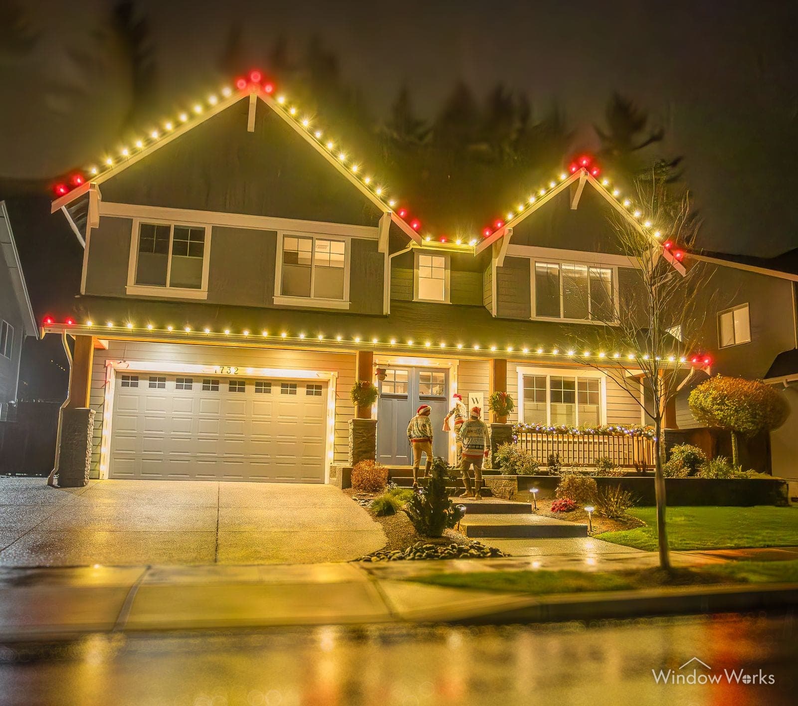 A grey two-story home in warm white with red bulbs marking the peaks and lit shrubs along the walk.