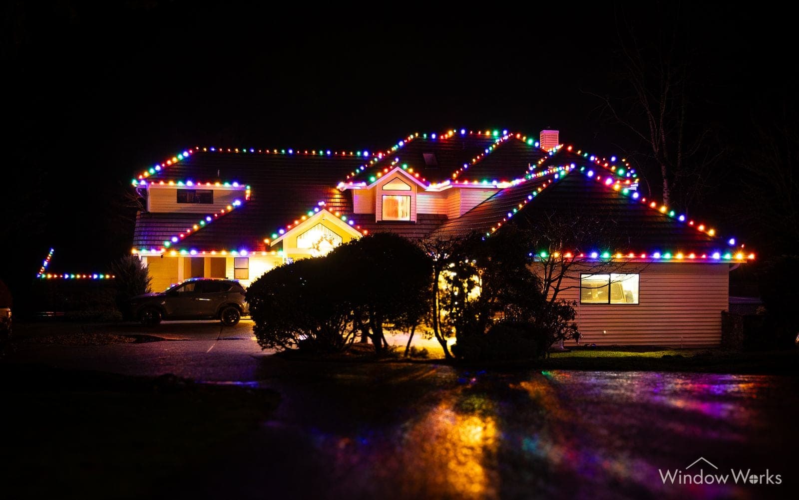 A ranch home in multicolor bulbs with the color reflected on a wet drive.