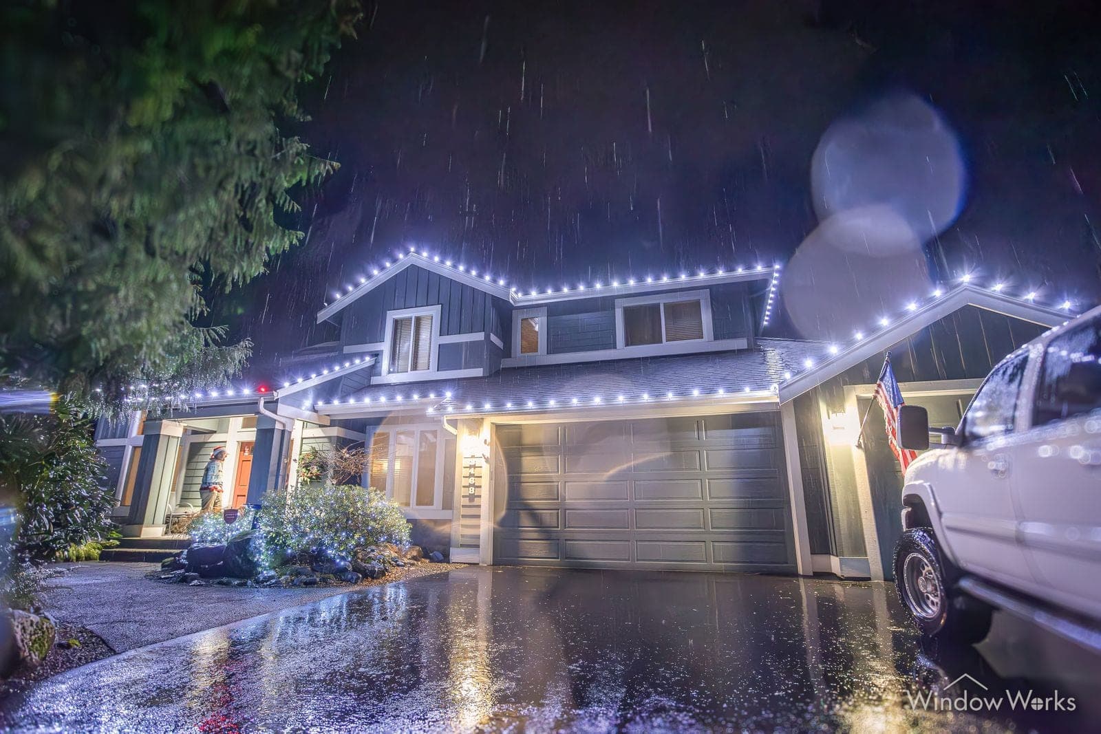 A split-level home outlined in warm white bulbs on a rainy night with a truck on the drive.