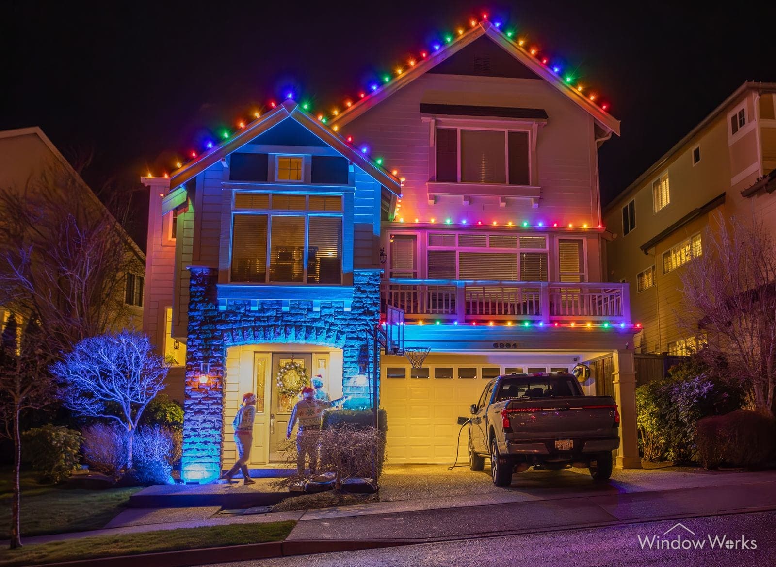 A three-story home lit blue, magenta and gold across its balconies with two people at the entry.