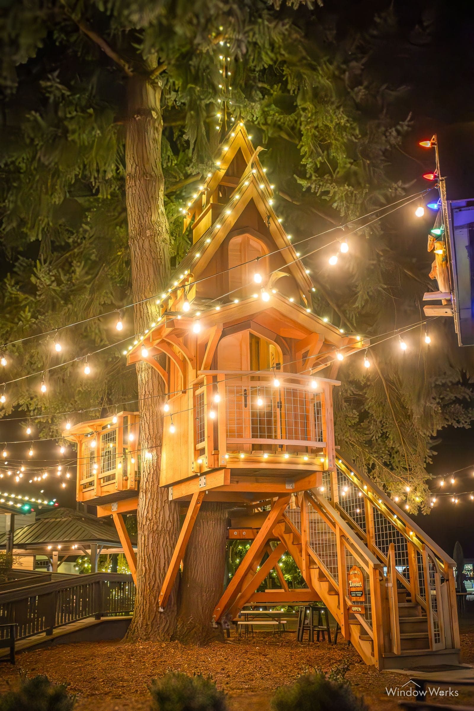 A two-story timber playhouse built into a fir, its roofline and stair rails run in warm white under a canopy of bulbs.