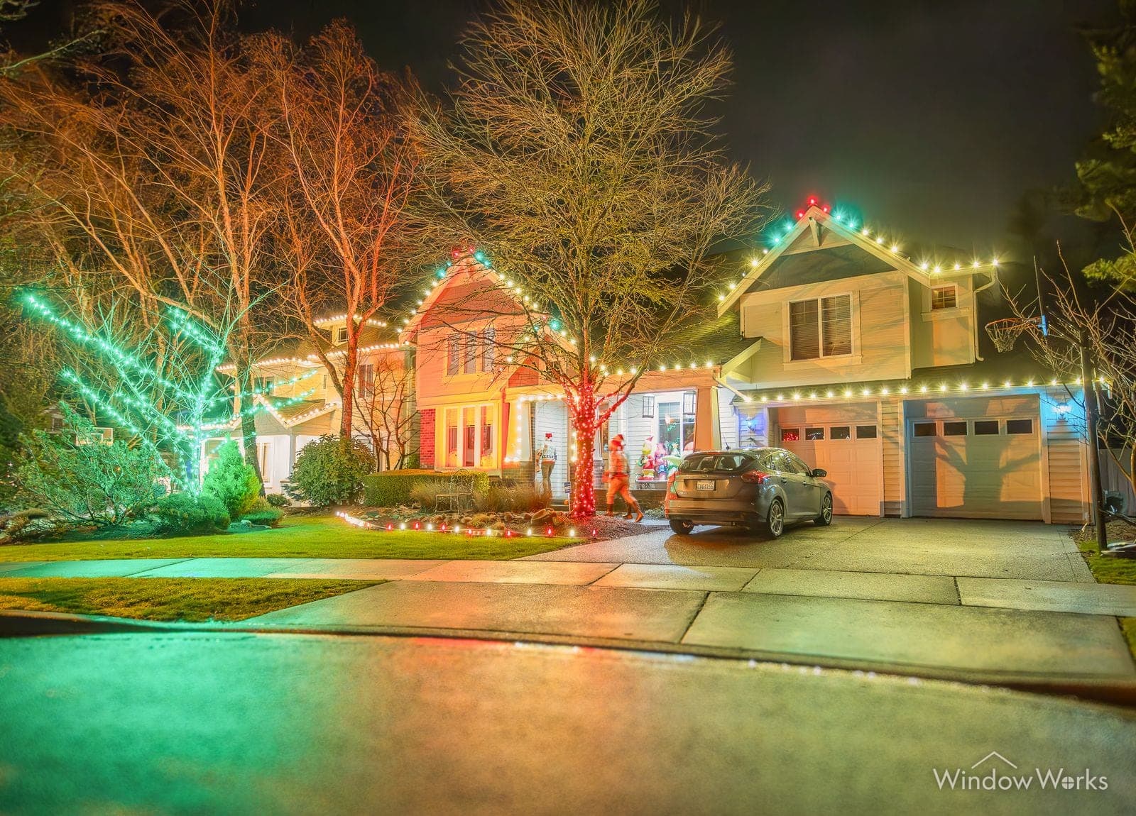 Two homes side by side lit in warm white and color, with trees uplit teal and red along the street.