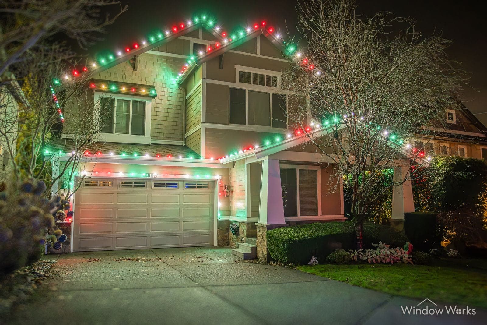 A two-story home in red and green bulbs with a bare tree wrapped in color beside the drive.