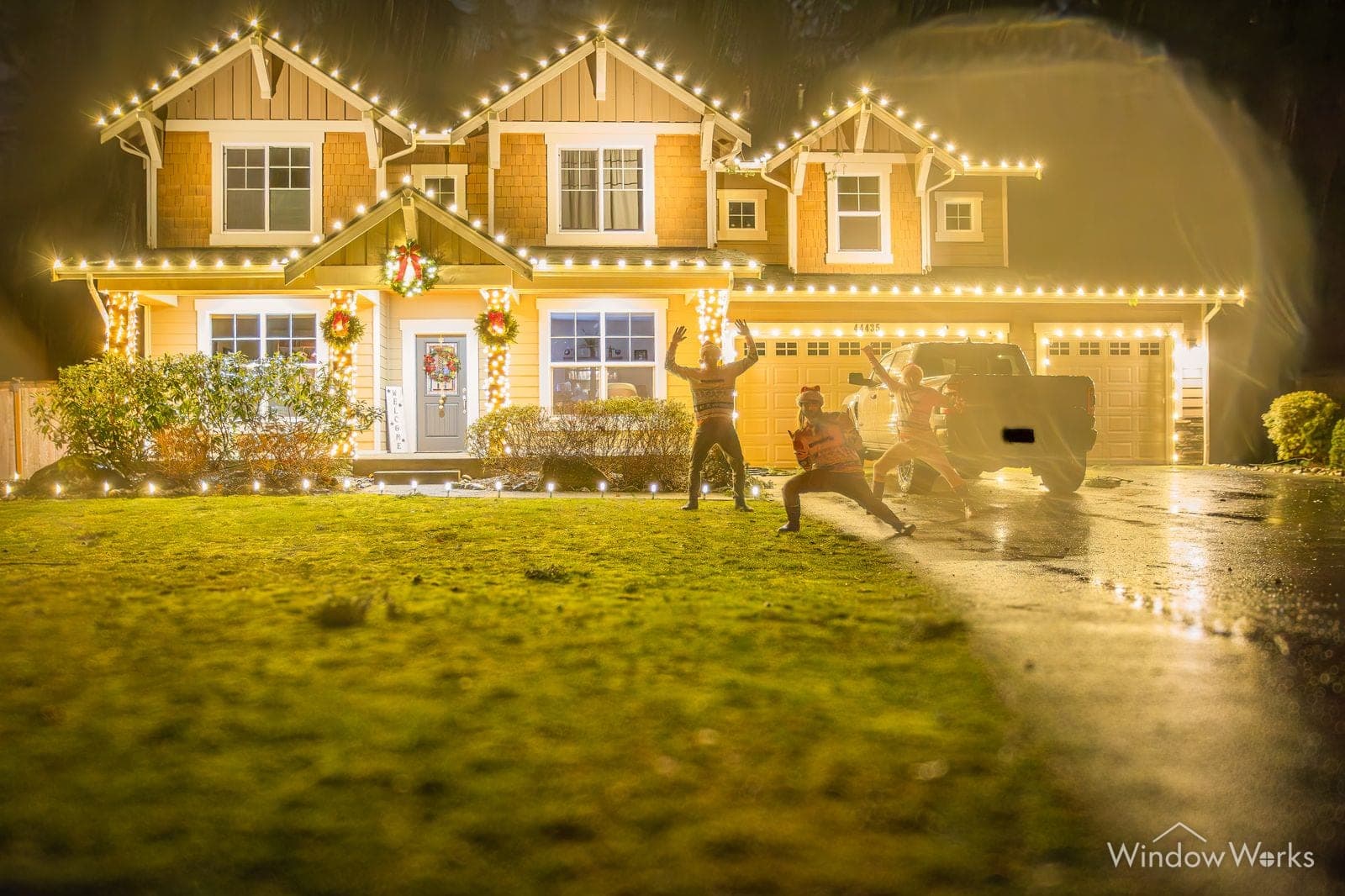 A home in warm white bulbs with wreaths and garland at the entry, two people crossing the wet drive.