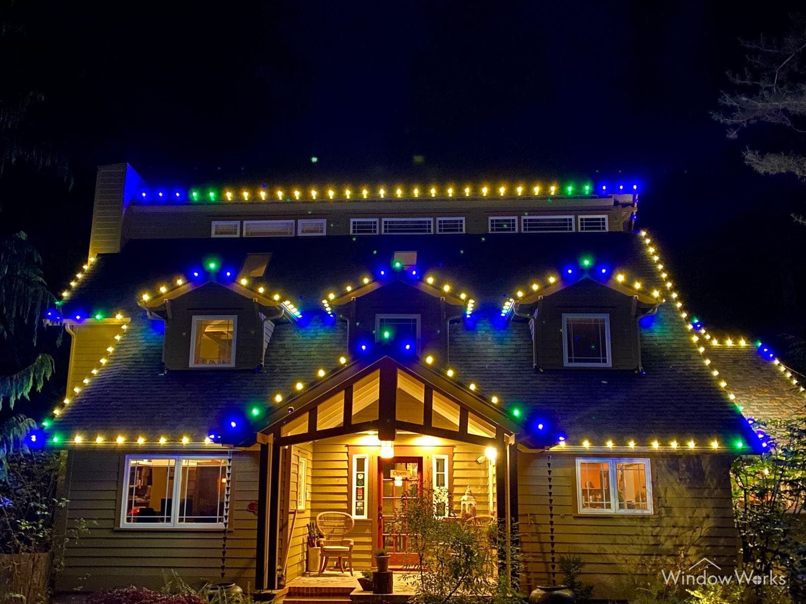 A craftsman home at night with blue and gold bulbs following every gable and the porch glowing below.
