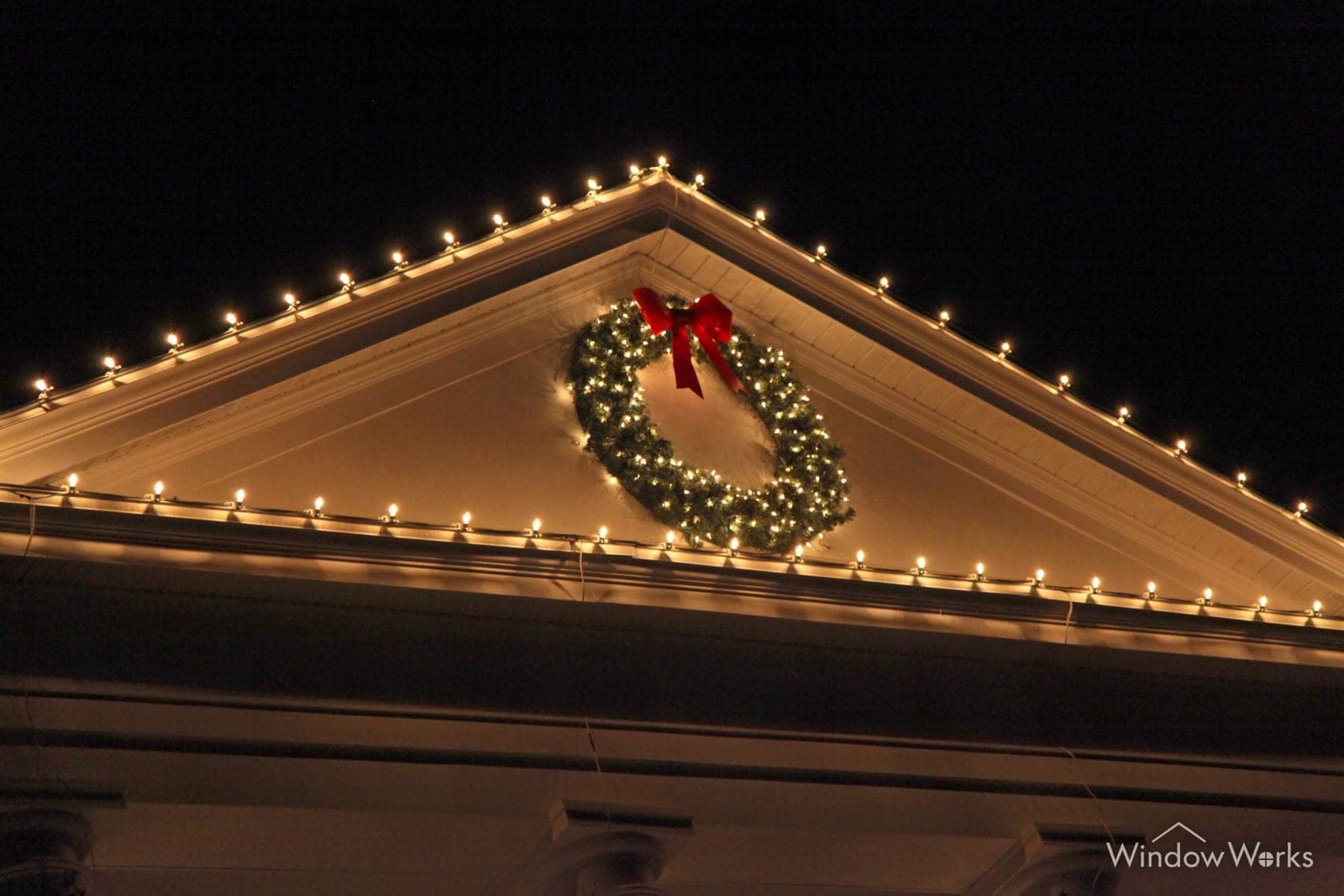 A gable outlined in warm white bulbs with a lit evergreen wreath and a red bow at its center.