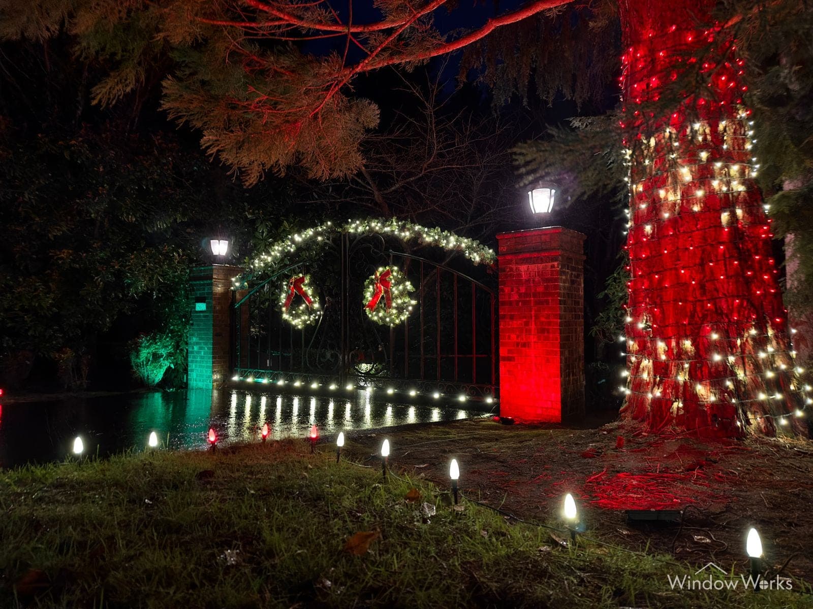 A lit entry gate with two wreaths, one stone column washed red and a fir wrapped in red beside it.