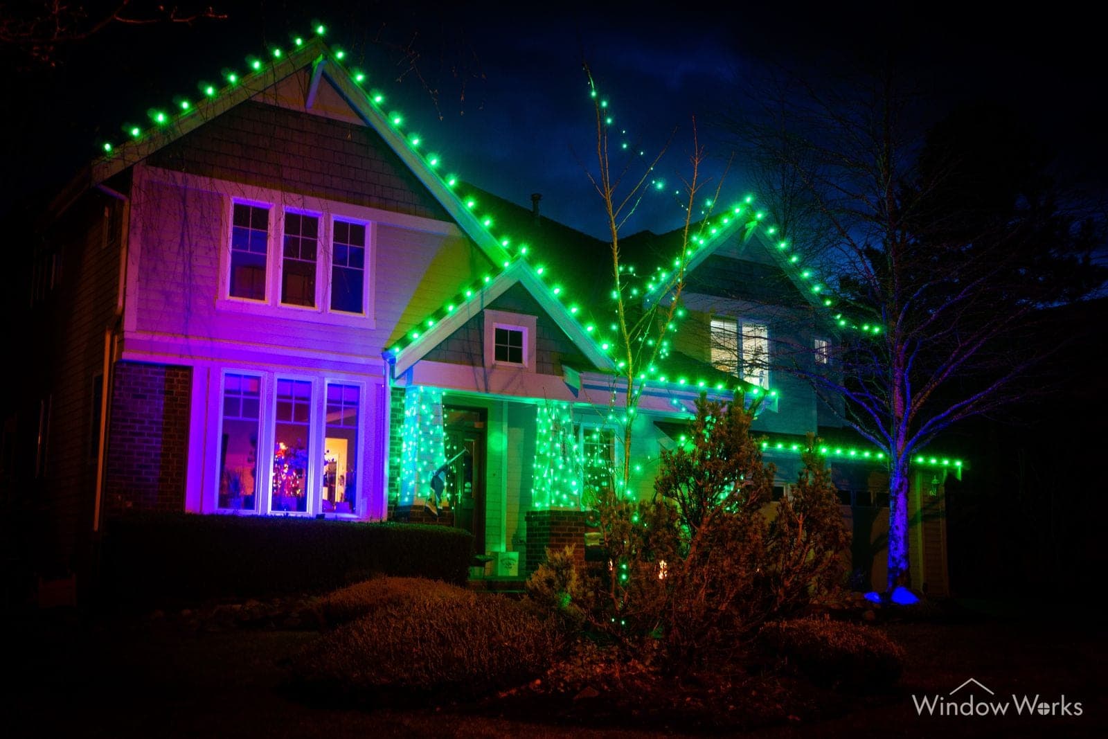A two-story home lit green along every roofline with magenta washing the walls and shrubs.
