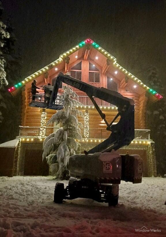 A boom lift parked in the snow below a log home, its roofline run in warm white with red and green at the peak.