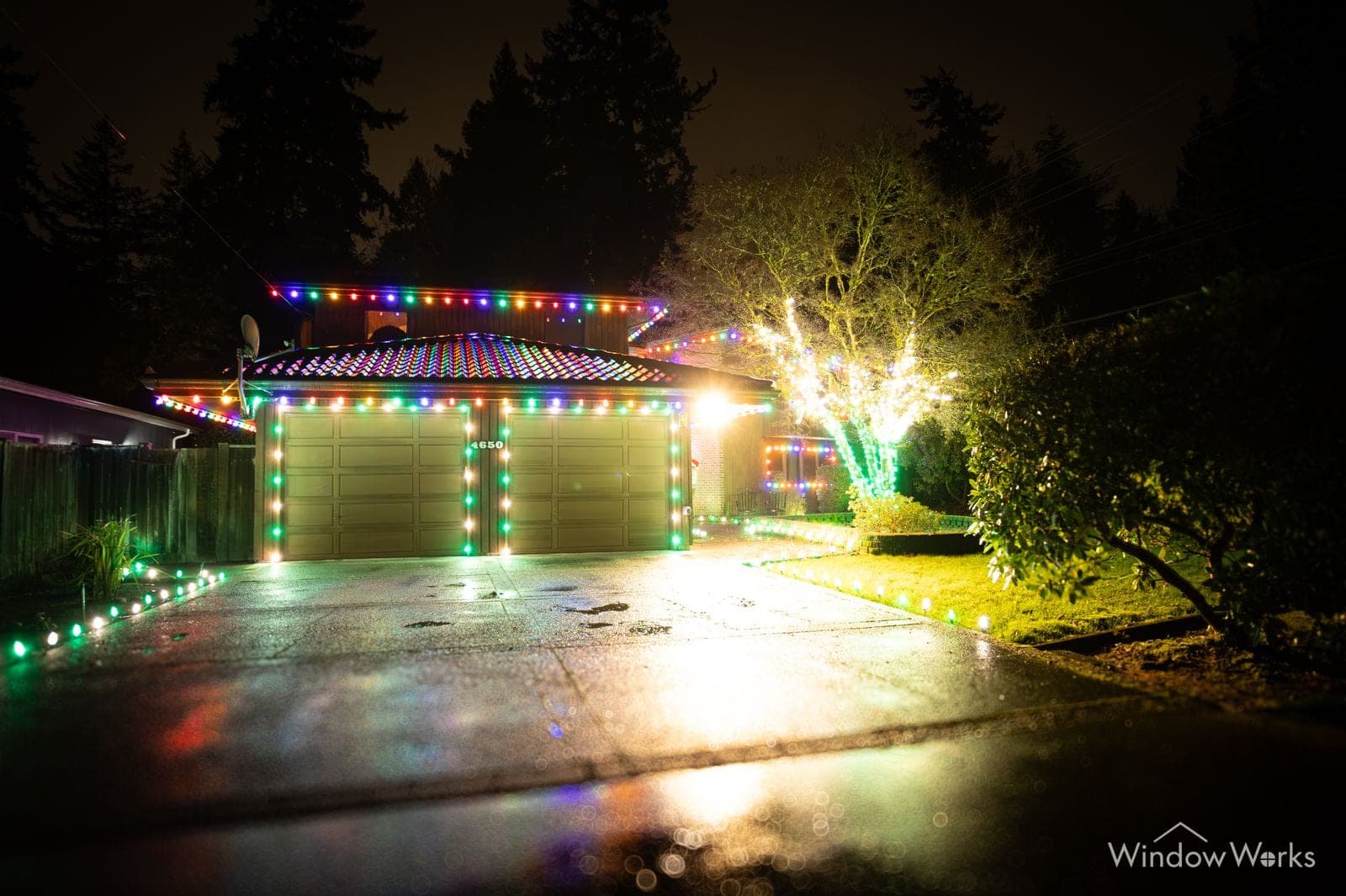 A tile-roofed home in multicolor bulbs with a brightly uplit tree at the corner and color running the drive.