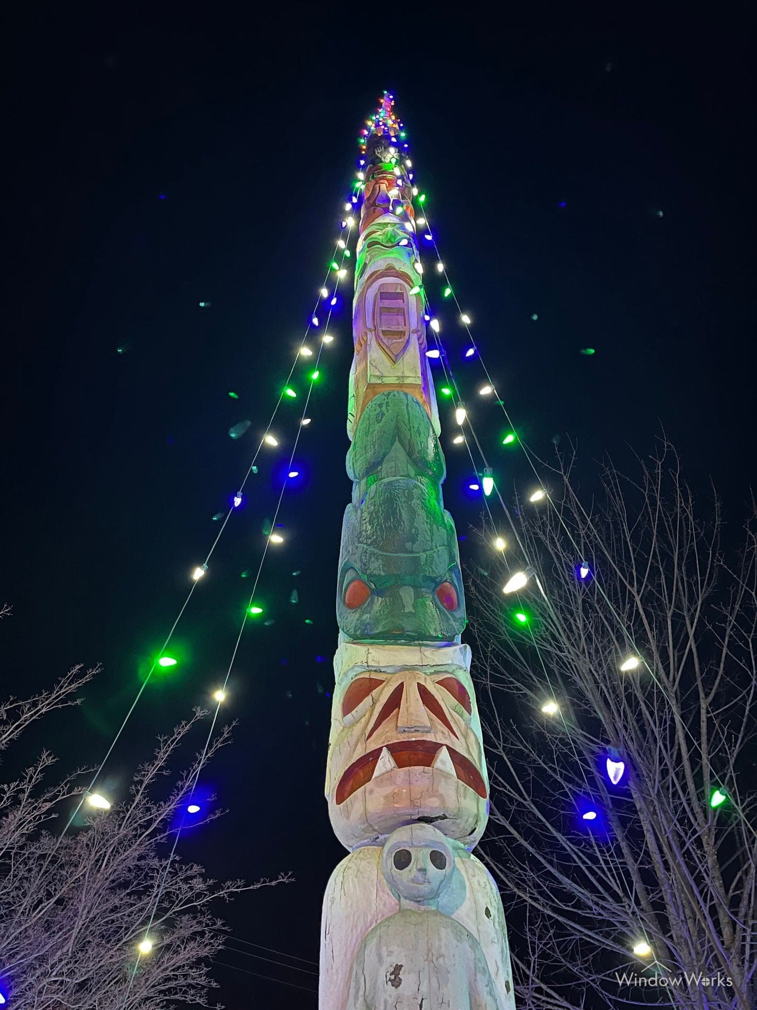 A tall carved totem pole strung with blue, green and white bulbs against a black sky.