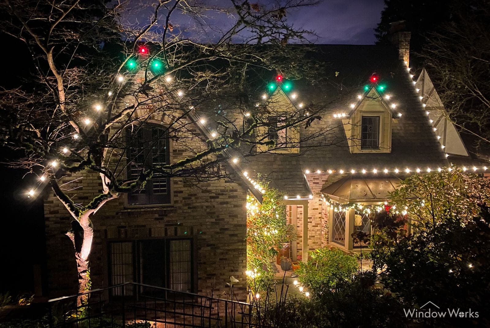 A tudor home in warm white bulbs with green and red at the peaks and a bare tree lit beside it.