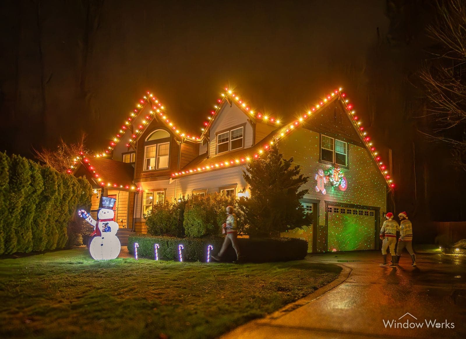 A home in warm white and red bulbs with a lit snowman on the lawn and crew walking the wet drive.