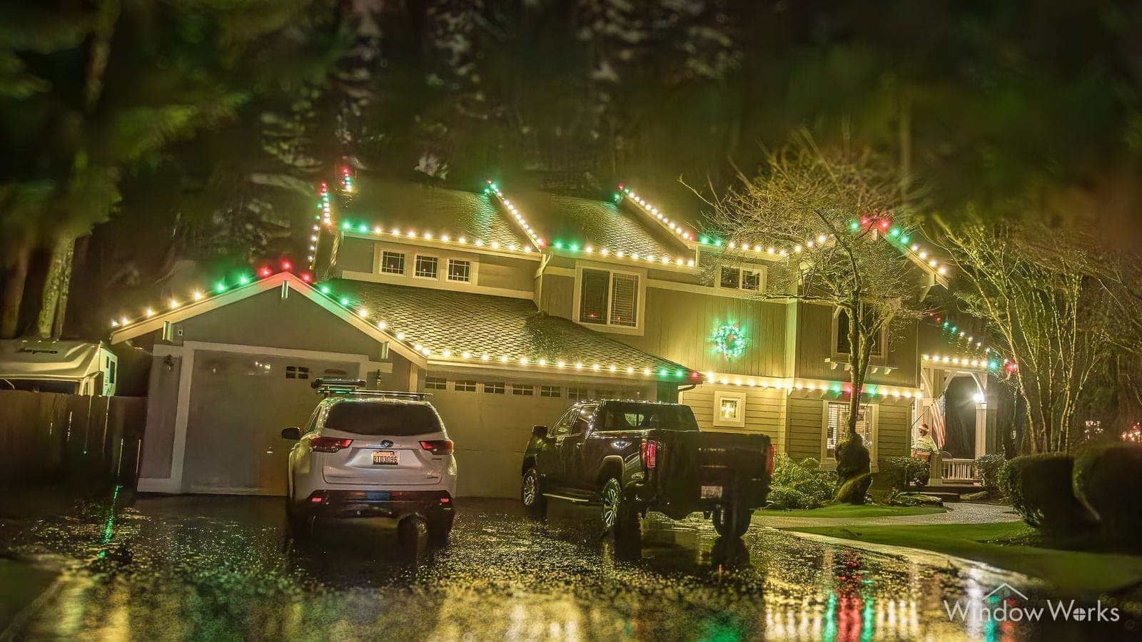 A home in warm white bulbs with green and red at the peaks, two vehicles on a wet drive and color reflected across it.
