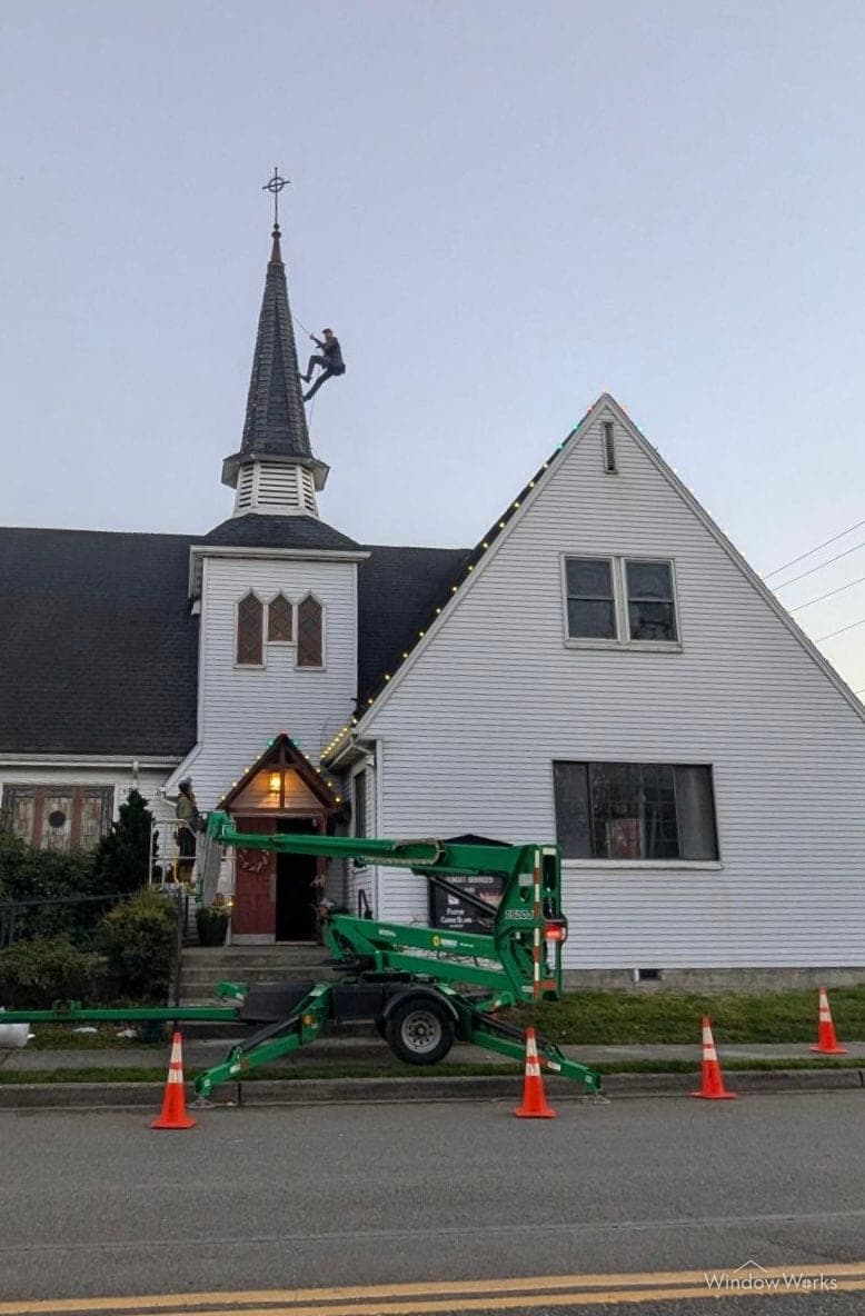 The lift parked at a white church in daylight, crew rigging the steeple for lights.