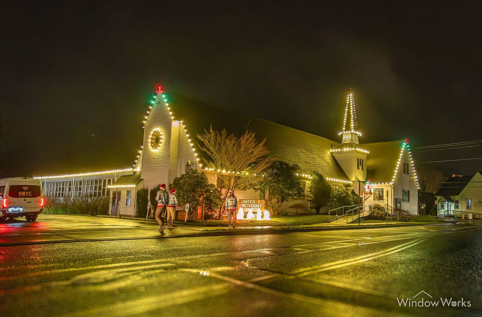 A church in warm white bulbs on a wet street at night, steeple lit beside it.