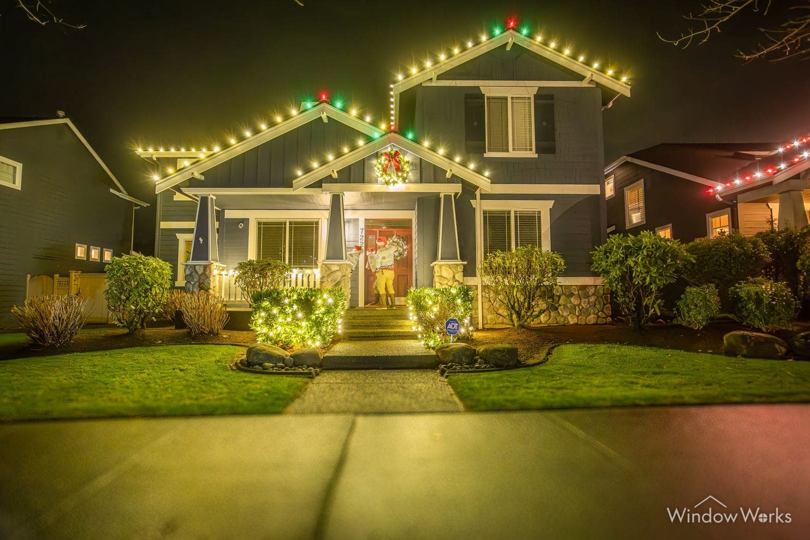 A craftsman home in warm white with red and green marking the gable peaks above lit beds.