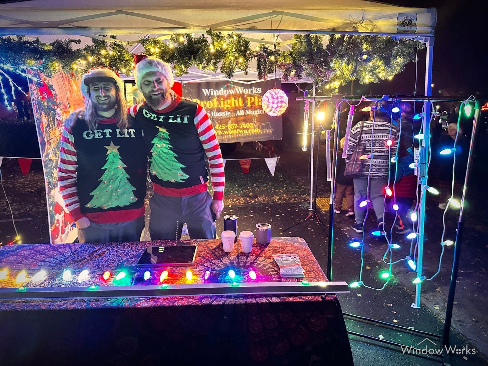 Two crew in matching holiday sweaters at the lit GlowWorks event booth, strung bulbs framing the table.