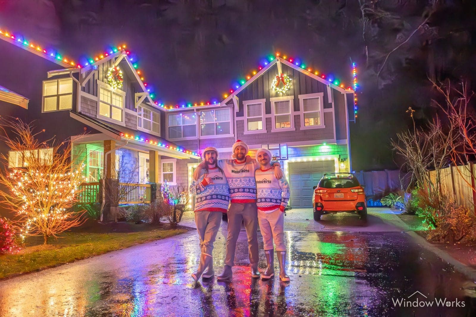 A family on the wet drive in front of their home lit in multicolor bulbs.