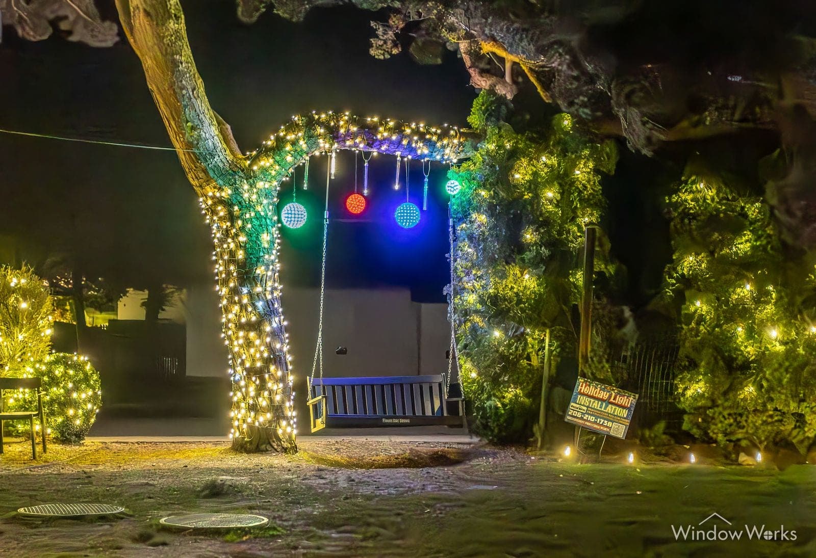 A garden swing hung with lit baubles under a bulb-strung tree, greenery lit behind.